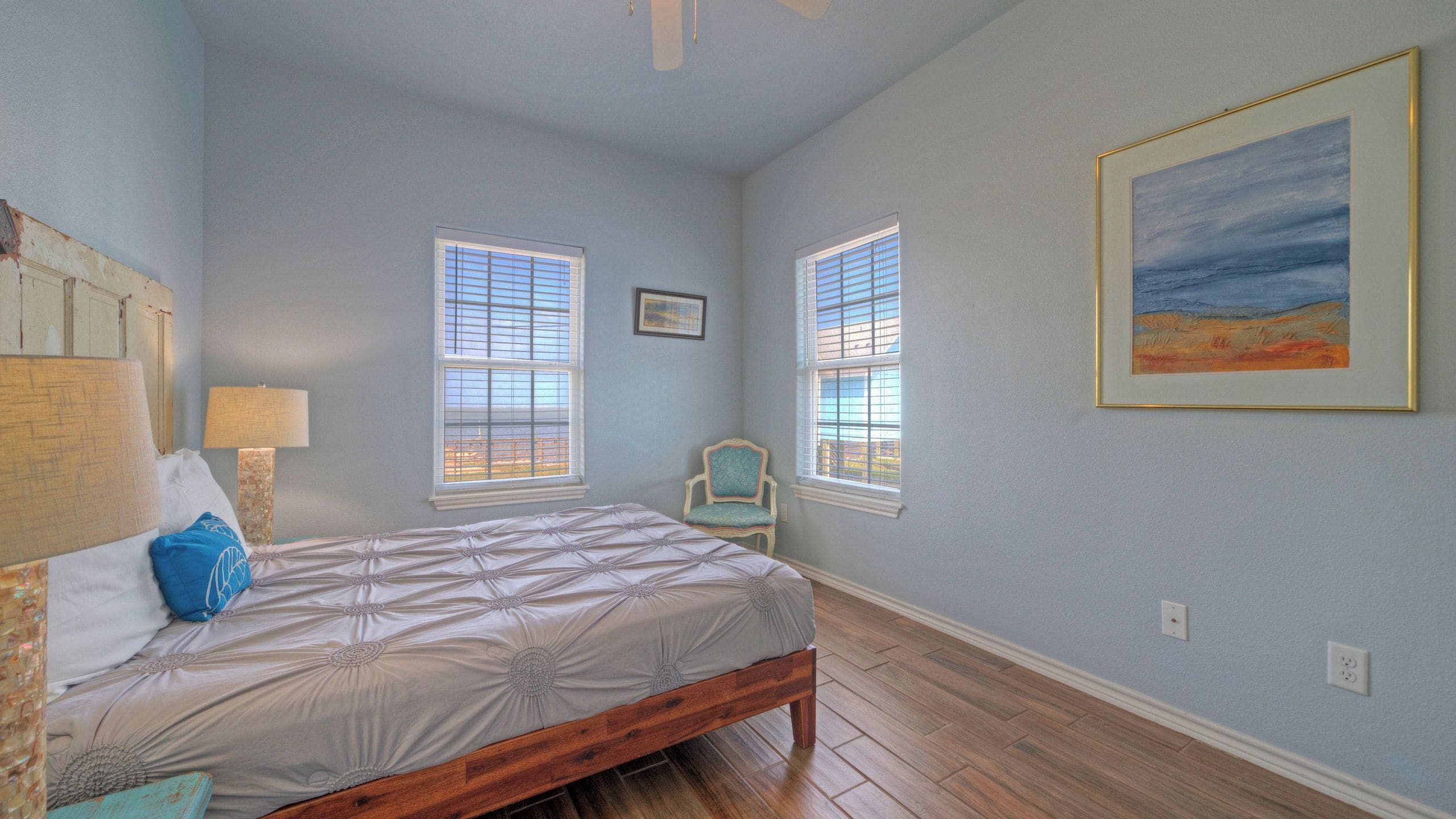 A bedroom with a bed, two windows, a chair, and a painting on the wall.