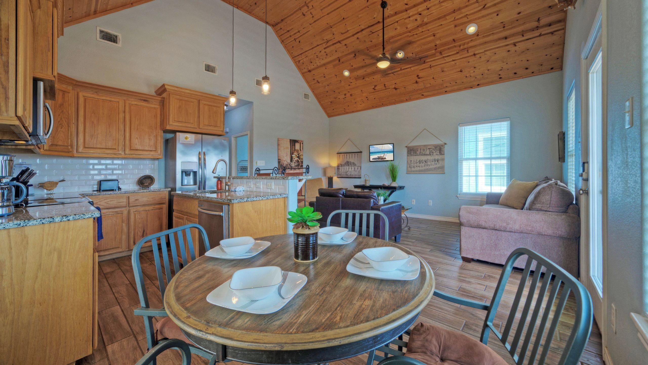 A spacious kitchen with wooden cabinets, a dining area with a round table, and a living room with a sofa and a window.