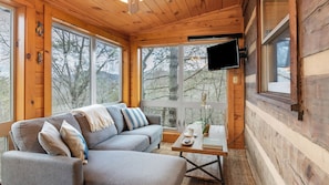 A cozy cabin interior with a sofa, a flat-screen TV, and large windows offering a view of trees. - Twilight Ridge by Avantstay Mountain Mansion, 20min to Pigeon Forge (Sevierville)