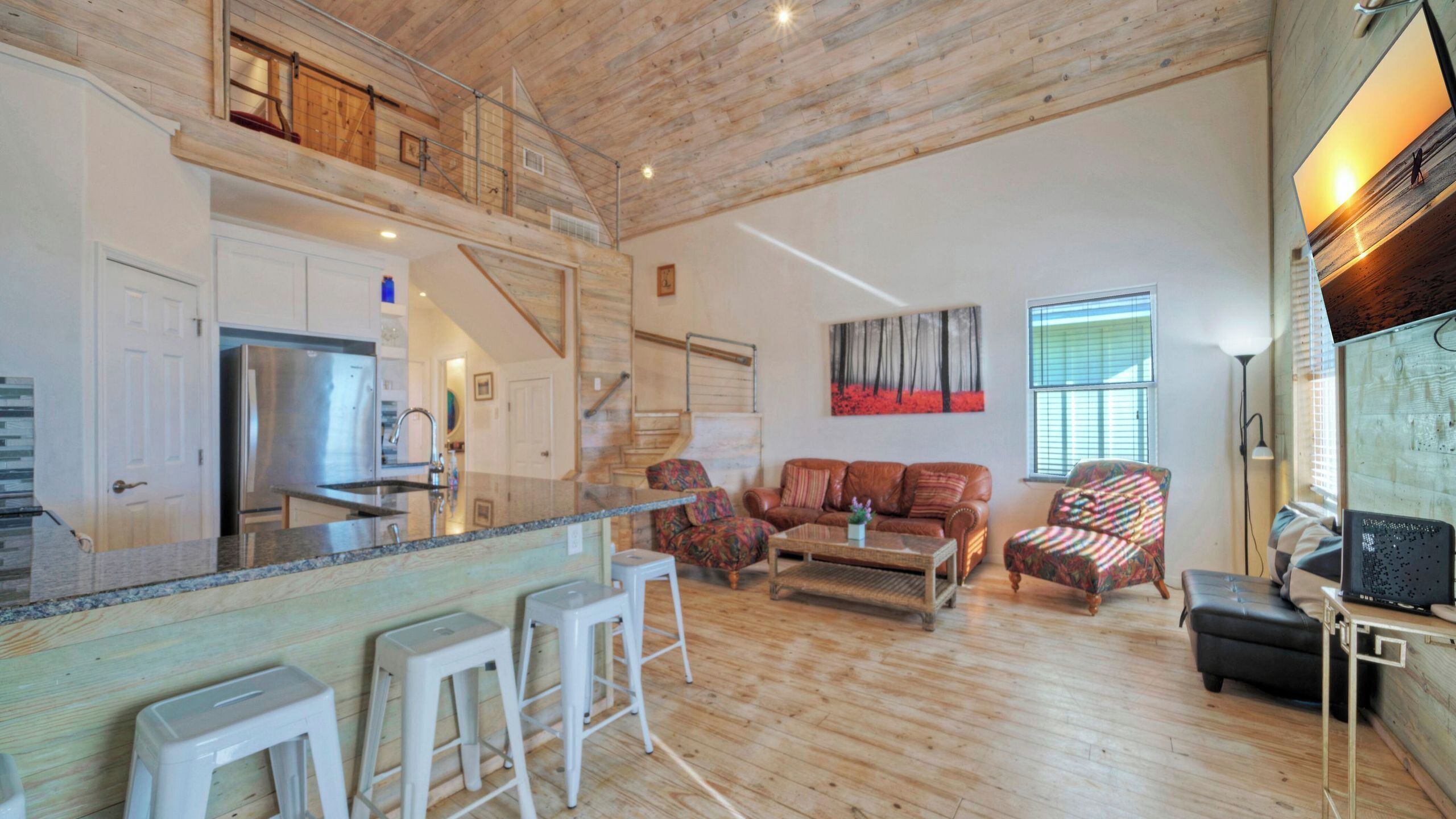 A spacious living area with a kitchen, wooden floors, a sofa set, and a large window with blinds.