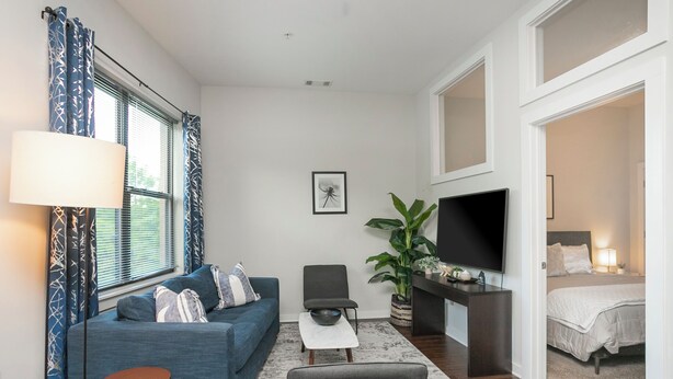 A modern living room with a blue sofa, a small table, a chair, a TV, and a bed in the adjacent room. - Music Row w Free Parking  Close to University (Nashville)