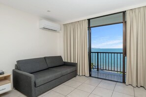 Een kamer met een grijze bank, een klein houten bijzettafeltje, een airconditioning en een schuifdeur die uitkomt op een balkon met uitzicht op zee.