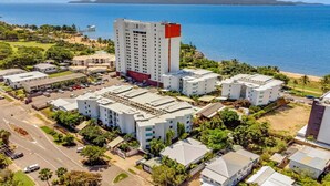 A coastal hotel complex with multiple buildings, a parking area, and a view of the ocean and mountains in the background.