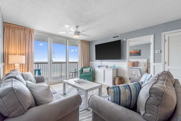 A living room with a sofa, armchairs, a coffee table, a TV, and a view of the ocean through large sliding glass doors.