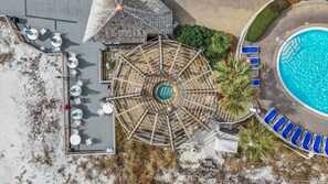 An aerial view of a resort with a central circular pool, surrounded by lounge chairs and a deck with tables and chairs.