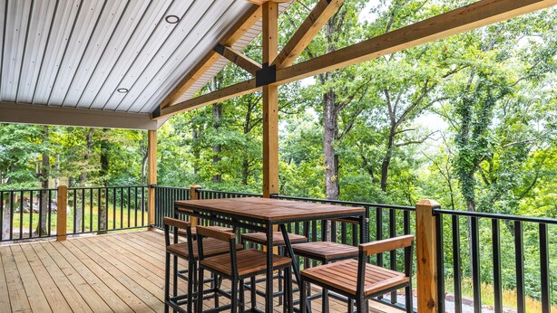 A covered outdoor deck with wooden tables and chairs, surrounded by a metal railing, overlooking a forested area.