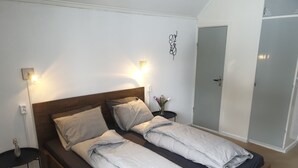 En dobbeltseng med træsengestolpe, to sengeborde, et lille bord med en blomstervase og en garderobe med spejl.