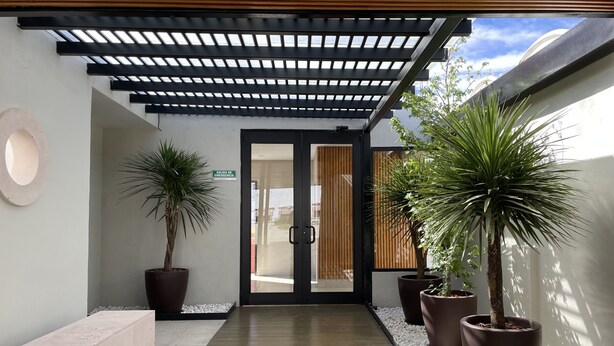 Um corredor com piso de madeira, plantas em vasos e uma porta com a inscrição "SALIDA DE EMERGENCIA".