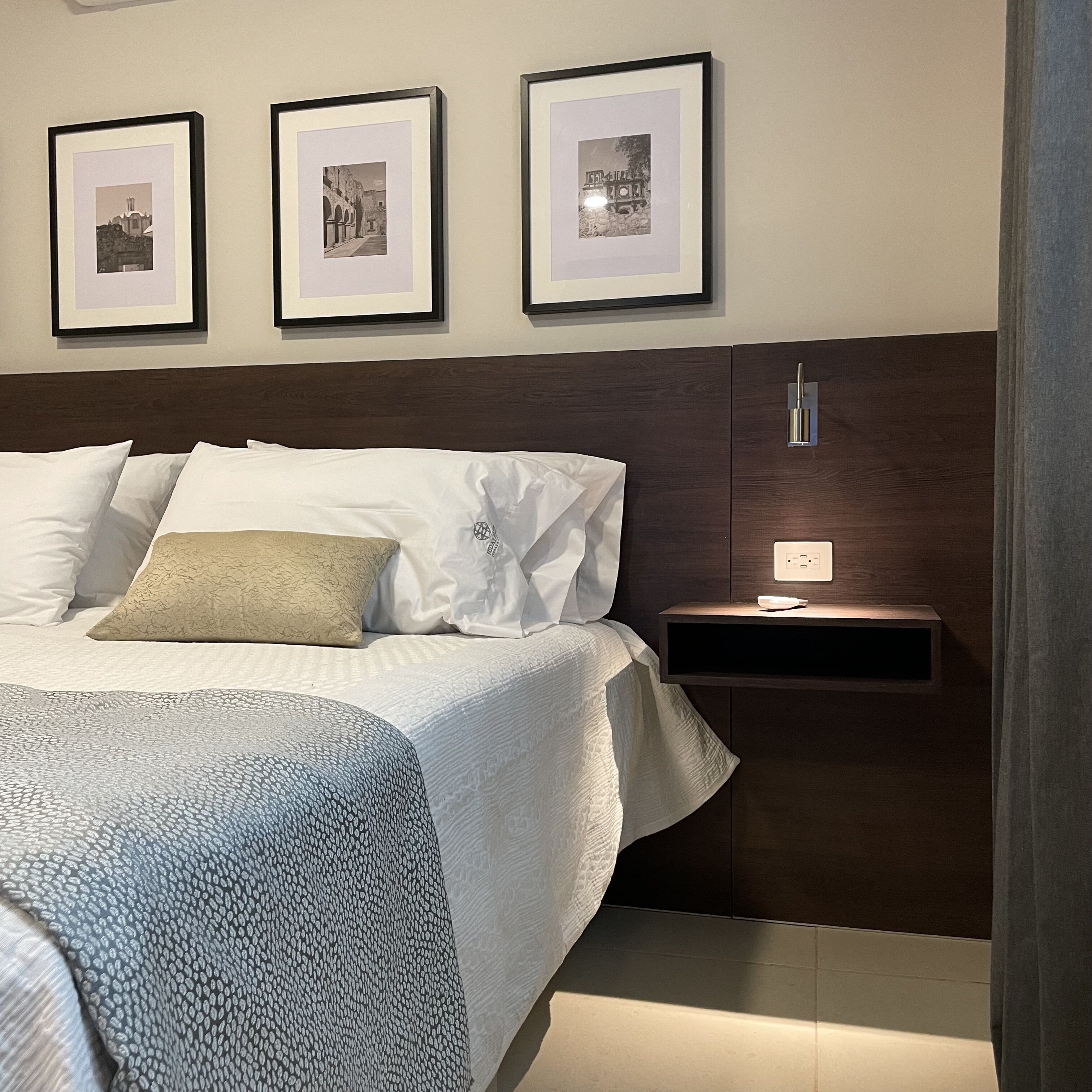 Una cama bien hecha con ropa de cama blanca y estampada, una mesita de noche con lámpara, y tres cuadros enmarcados en la pared sobre la cama.