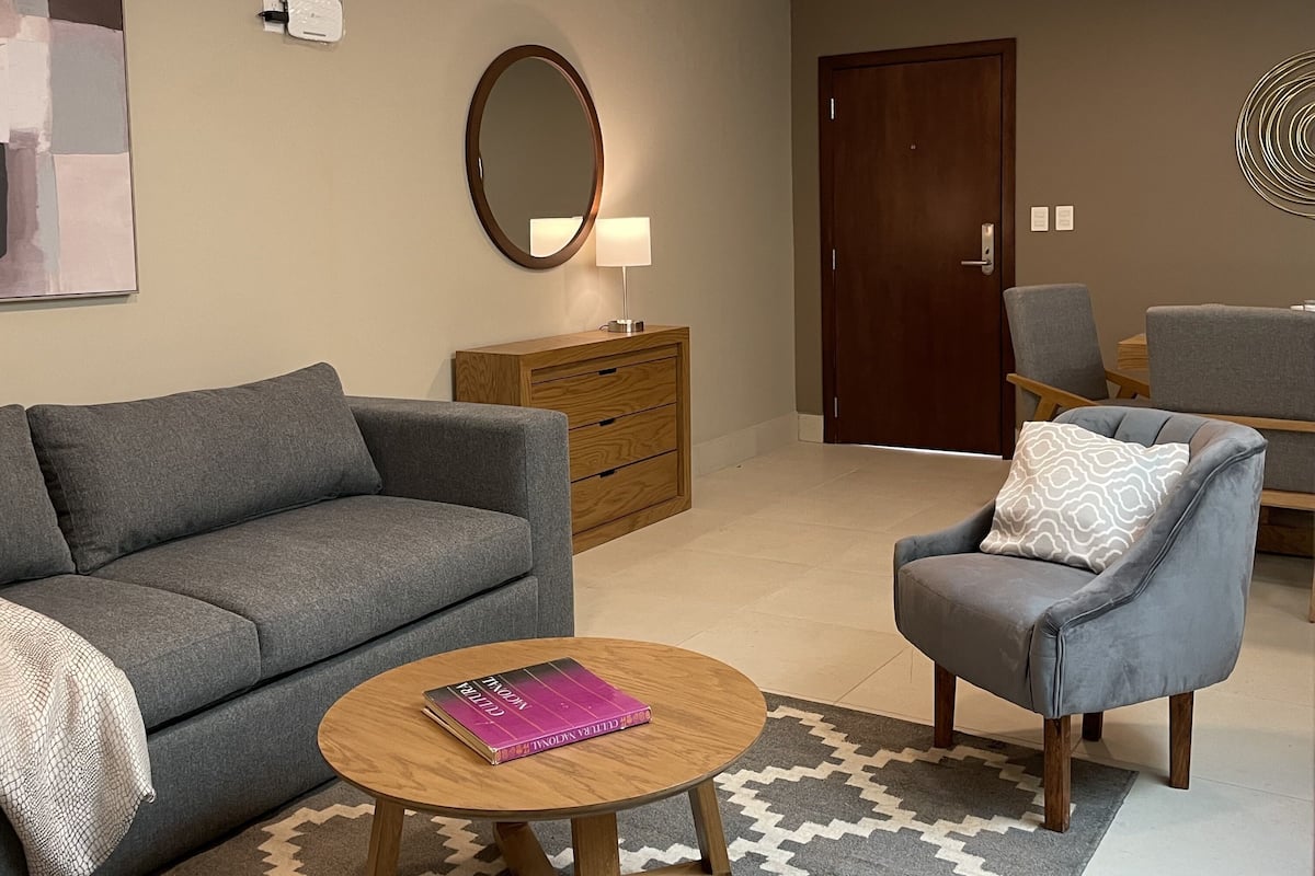A living room with a grey sofa, a round wooden coffee table with a book, a grey armchair, a wooden side table with a lamp, a large mirror, and a patterned rug.
