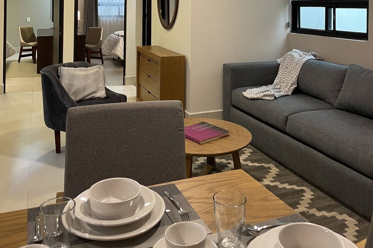 A modern hotel room with a dining area, a sofa, a mirror, and a view of the outside.