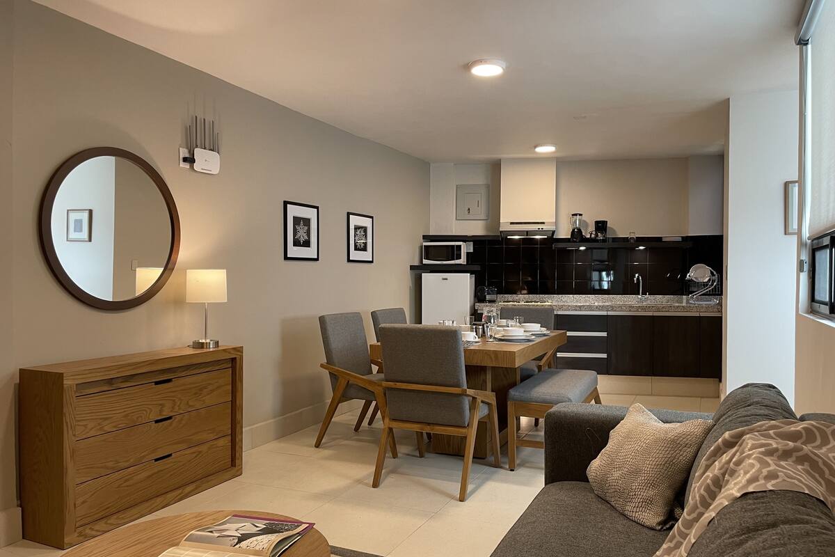 A modern living room with a sofa, a wooden side table, a dining area with chairs, and a kitchen in the background.