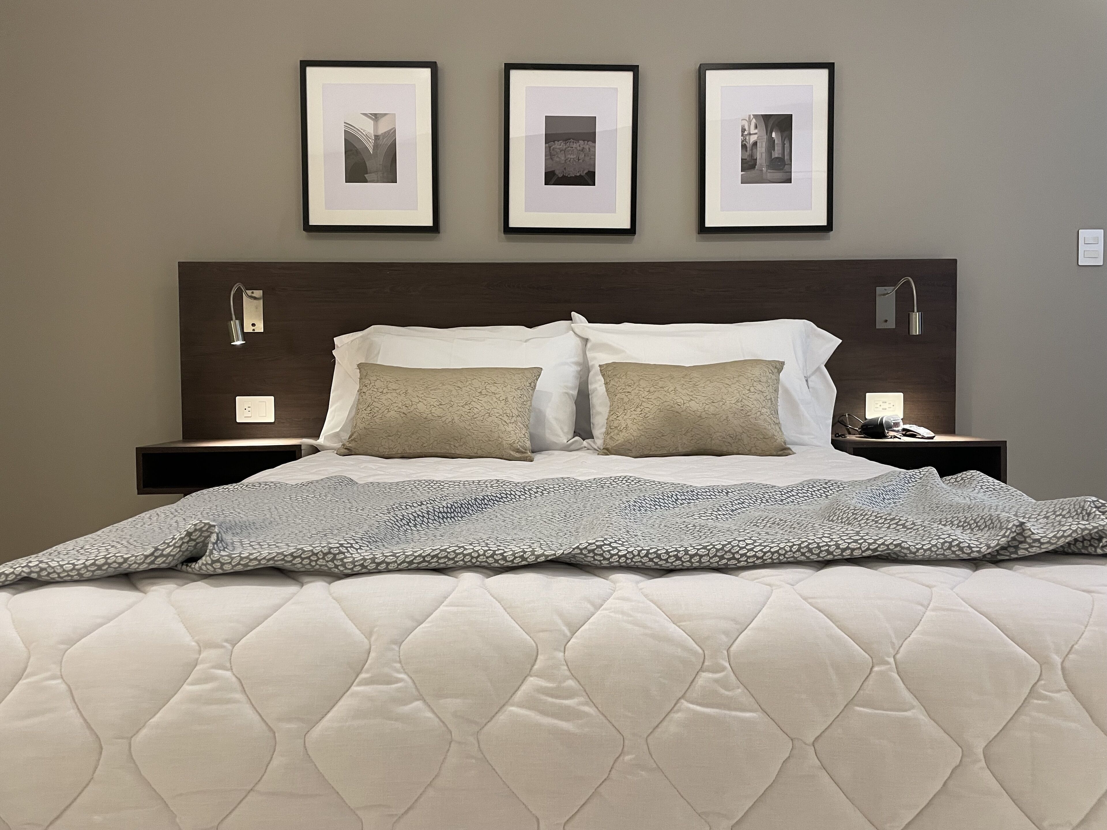 Una cama bien hecha con cubrecolcha acolchado, dos mesitas de noche con lámparas, y tres cuadros enmarcados en la pared sobre la cama.