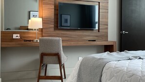 Quarto de hotel moderno com uma grande TV de tela plana presa a uma parede revestida de painel de madeira, uma escrivaninha com abajur, uma poltrona confortável e uma cama bem arrumada com um cobertor estampado.