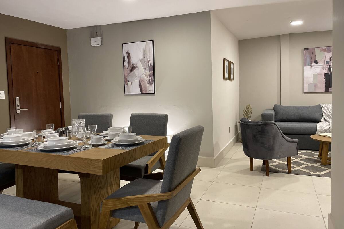 A dining area with a wooden table set for four, surrounded by grey upholstered chairs. There are two framed abstract paintings on the walls, a grey sofa, and a small round table with a plant.