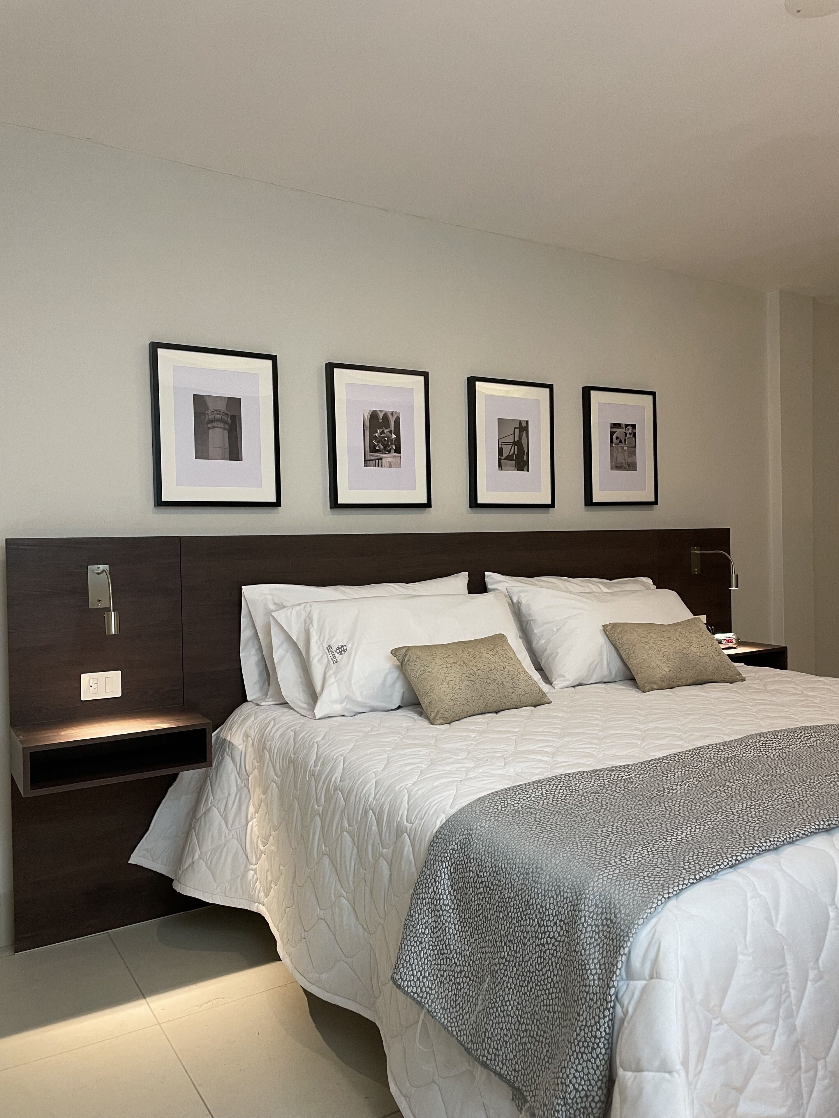 A neatly arranged bedroom with a large bed, a dark wooden headboard, and a nightstand with a lamp. There are four framed pictures on the wall above the bed.