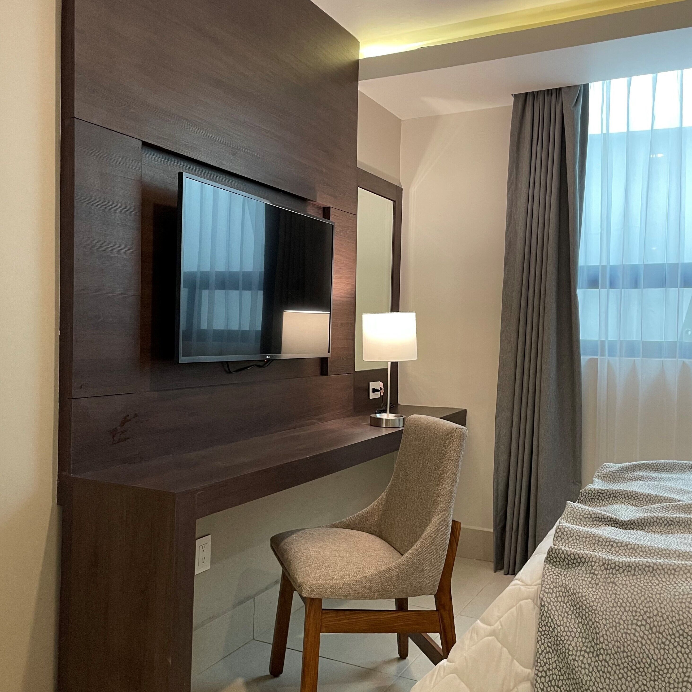 A hotel room with a flat-screen TV mounted on a wooden panel, a bedside lamp, a chair, and a bed with a patterned bedspread.