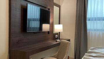 A hotel room with a flat-screen TV mounted on a wooden panel, a bedside lamp, a chair, and a bed with a patterned bedspread.