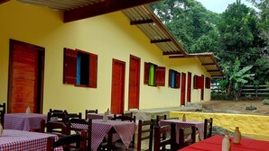 An outdoor dining area with tables and chairs, a yellow building with red and green doors, and a view of greenery in the background. - Pousada Sítio Monte Sinai (São José do Barreiro)