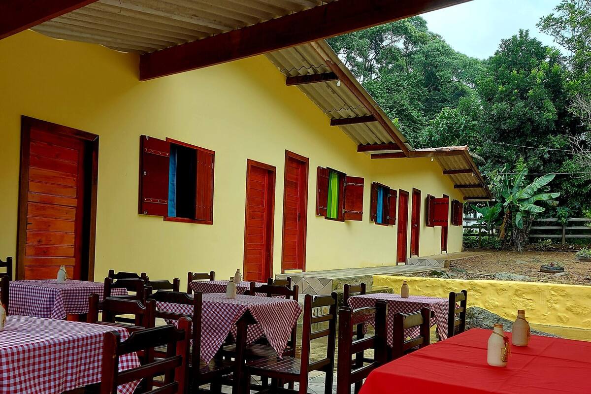Un área de comedor al aire libre con mesas y sillas, un edificio amarillo con puertas rojas y verdes, y vistas a la vegetación en el fondo.