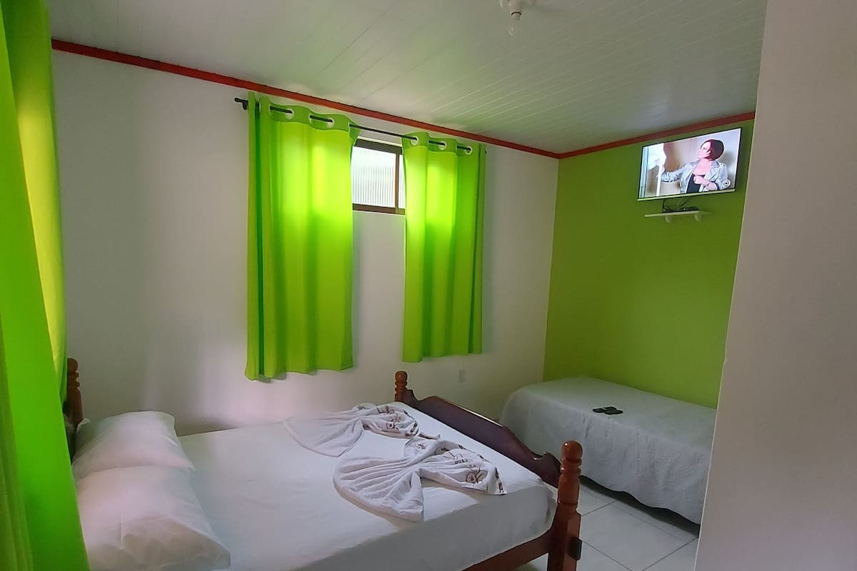Una habitación con una cama individual, una cama más pequeña, cortinas verdes, un televisor en la pared y una ventana con persianas.
