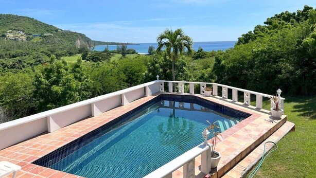 Una piscina rectangular amb vora enrajolada, envoltada d'una barana blanca, amb un fons de vegetació exuberant i una vista llunyana del mar.