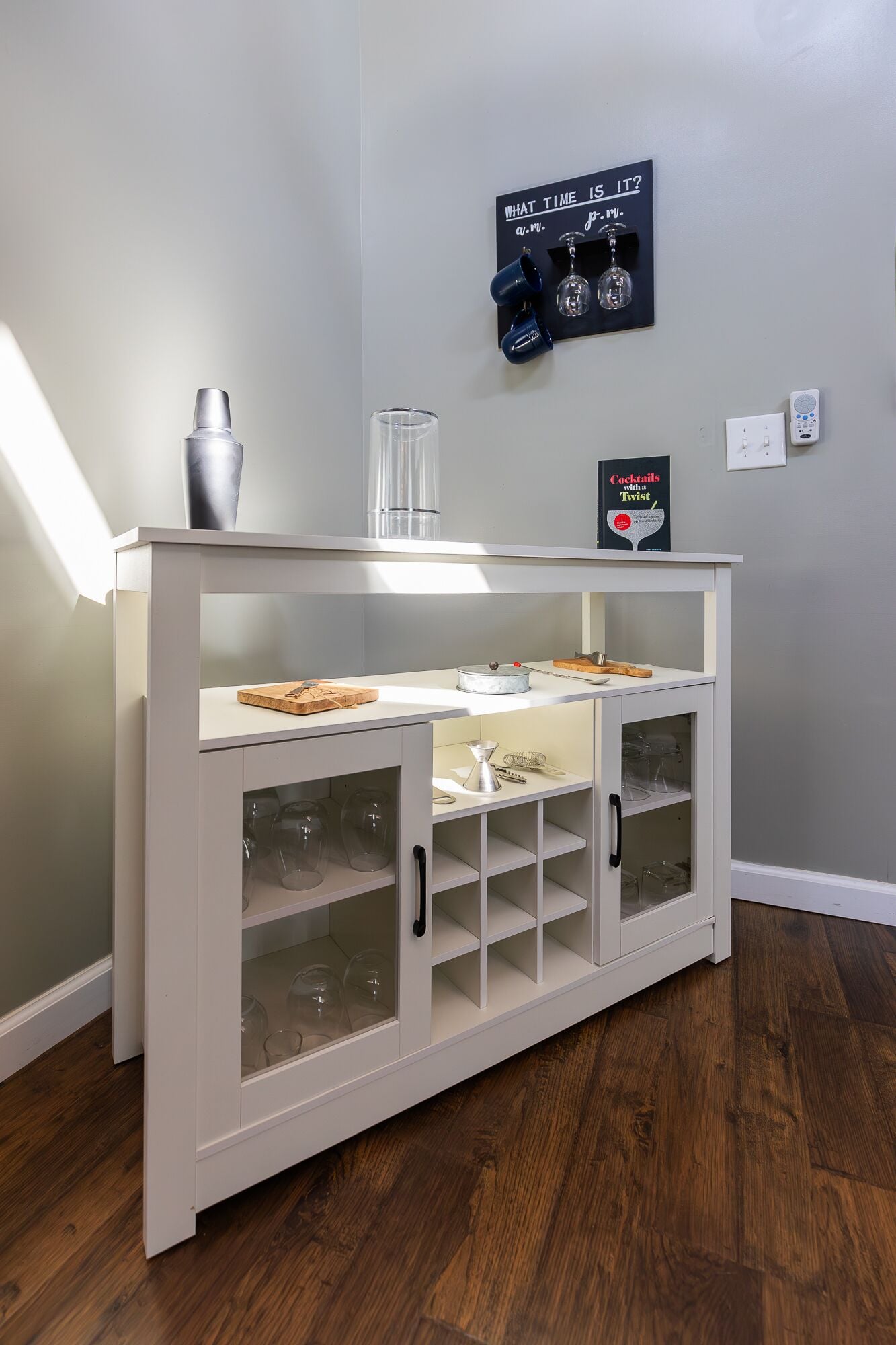Ein weißer Barwagen mit einer Glasvitrine, einem Metallsieb und einem Cocktail-Rezeptbuch an der Wand.