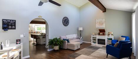 A spacious living area with a ceiling fan, a kitchen in the background, a large clock, and a blue armchair.
