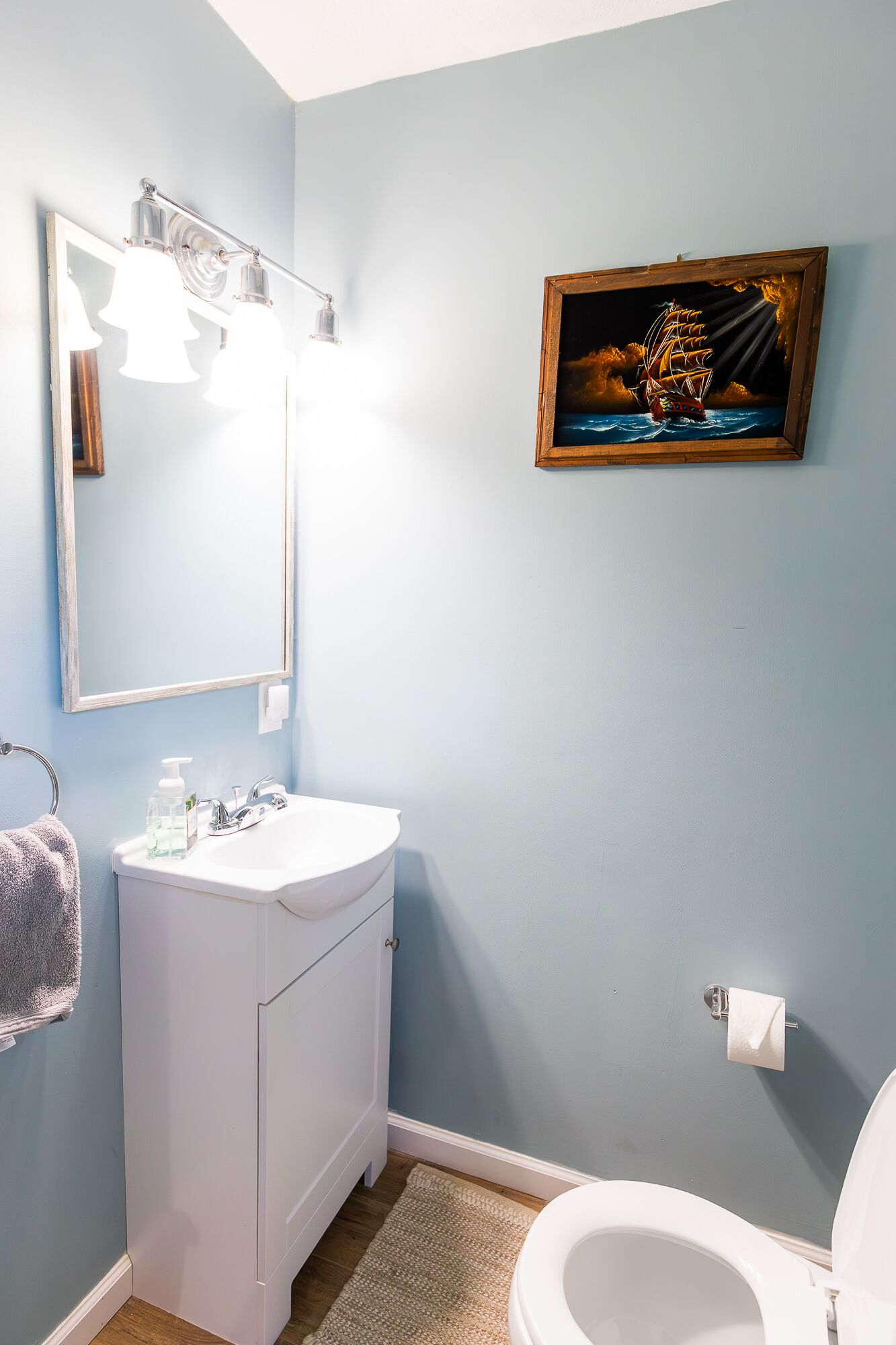 Une salle de bain avec un meuble-lavabo blanc, un miroir, un tableau représentant un navire et des toilettes.