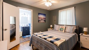 Une chambre à coucher comprenant un lit, des tables de chevet, une commode, un ventilateur de plafond, une fenêtre avec des rideaux et un tableau accroché au mur.