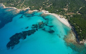 An aerial view of a coastal area with clear turquoise waters, a sandy beach, and surrounding greenery.