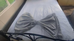 Yhden hengen sänky, jossa on metallinen runko, valkoiset vuodevaatteet ja pääty, jossa on hylly yläosassa. Toisella puolella on ikkuna ja toisella pieni pöytä, jossa on lamppu.