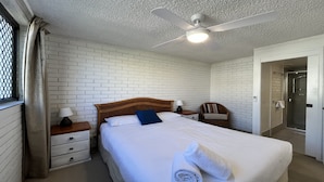 Ein Hotelzimmer mit weißer Backsteinwand, einem hölzernen Bett mit weißer Bettwäsche, einem Deckenventilator, einem Nachttisch mit Lampe, einem Sessel und einem Badezimmer mit Duschkabine.