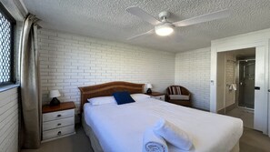 A hotel room with a white brick wall, a wooden bed with white bedding, a ceiling fan, a nightstand with a lamp, a chair, and a bathroom with a glass shower. - Edgecliffe 3 Kings Beach Unit With Ocean Views (Caloundra)