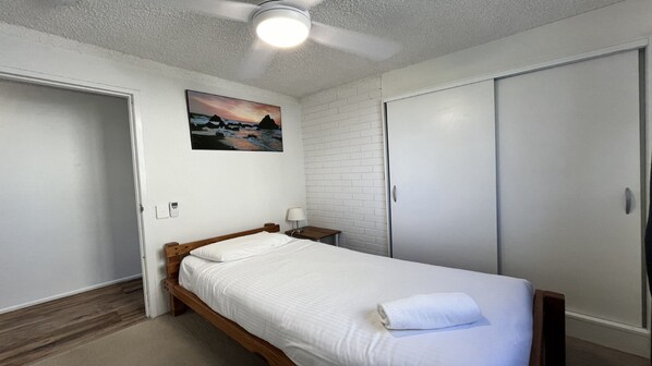 Un dormitorio con cama, mesita de noche, lámpara de pared, ventilador de techo e imagen de un paisaje.