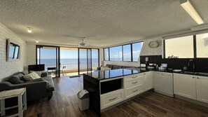 Una cocina moderna con una isla grande, un comedor con vista al océano y una sala de estar con sofá y televisor.
