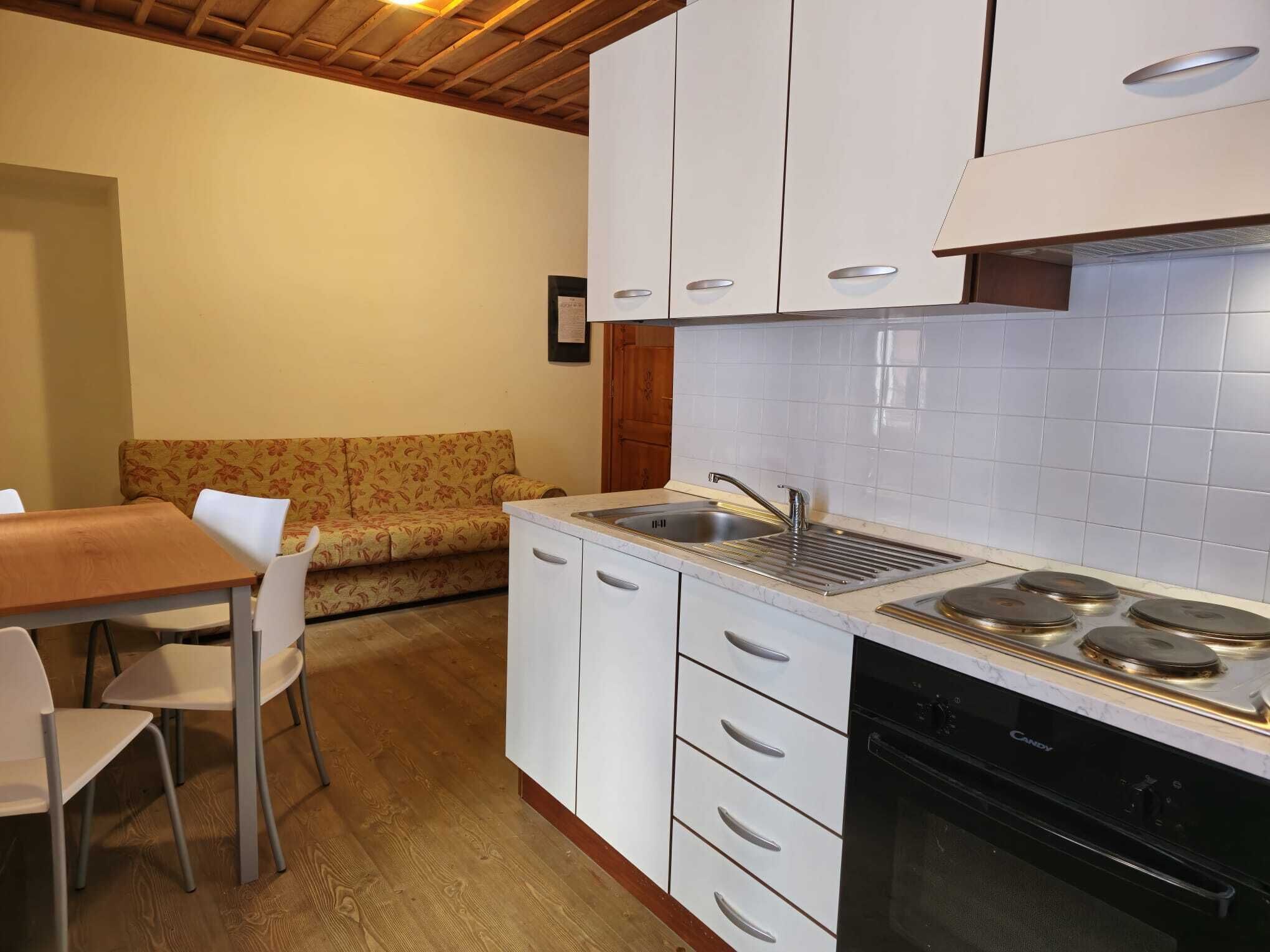 Een kleine keuken met witte kastjes, een inbouwfornuis en een spoelbak. Er staat een houten eettafel met stoelen en een bloemenbank.