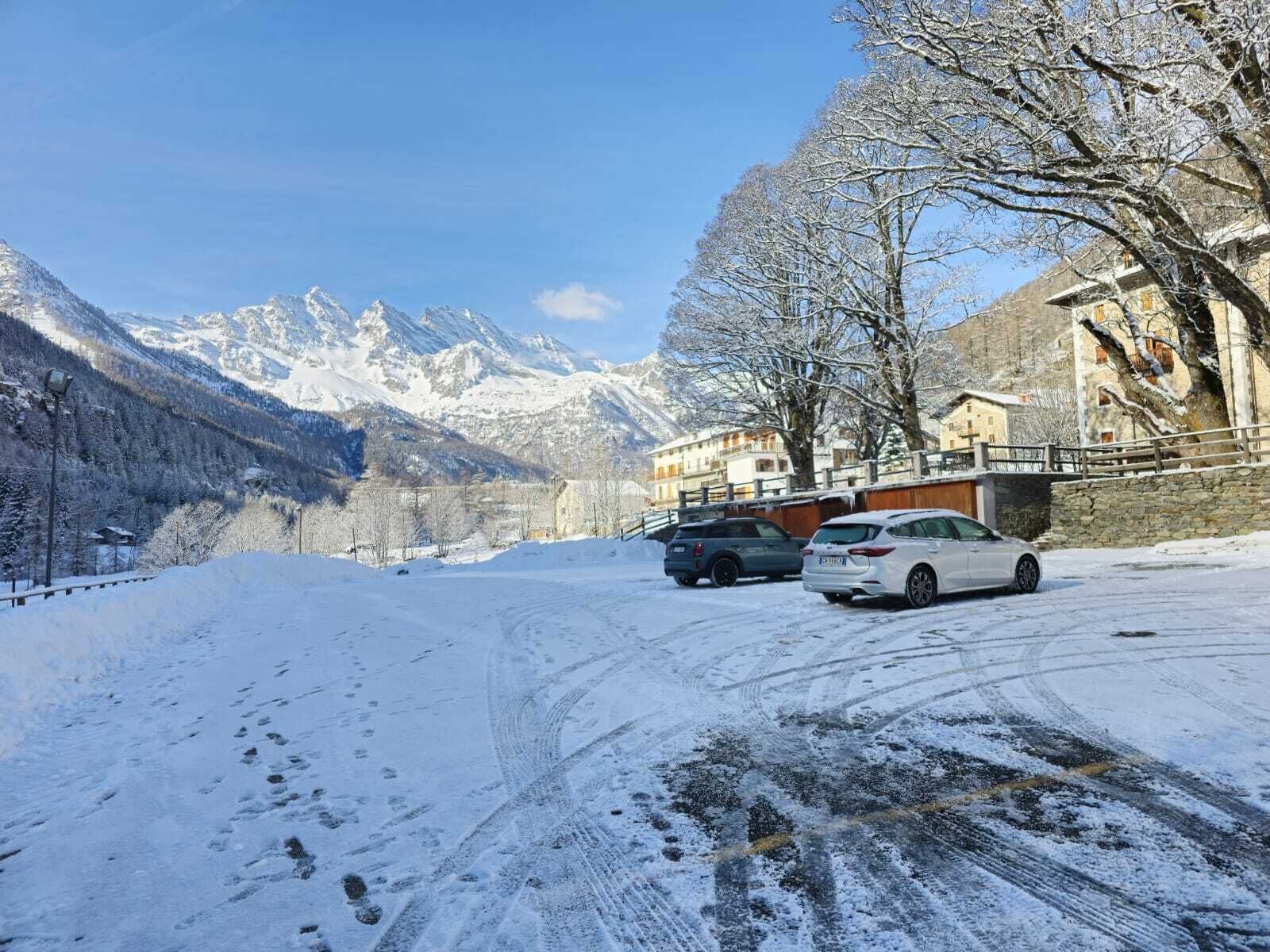 Eine verschneite Bergstraße mit parkenden Autos, ein Steinhaus mit Balkon und schneebedeckten Bäumen.