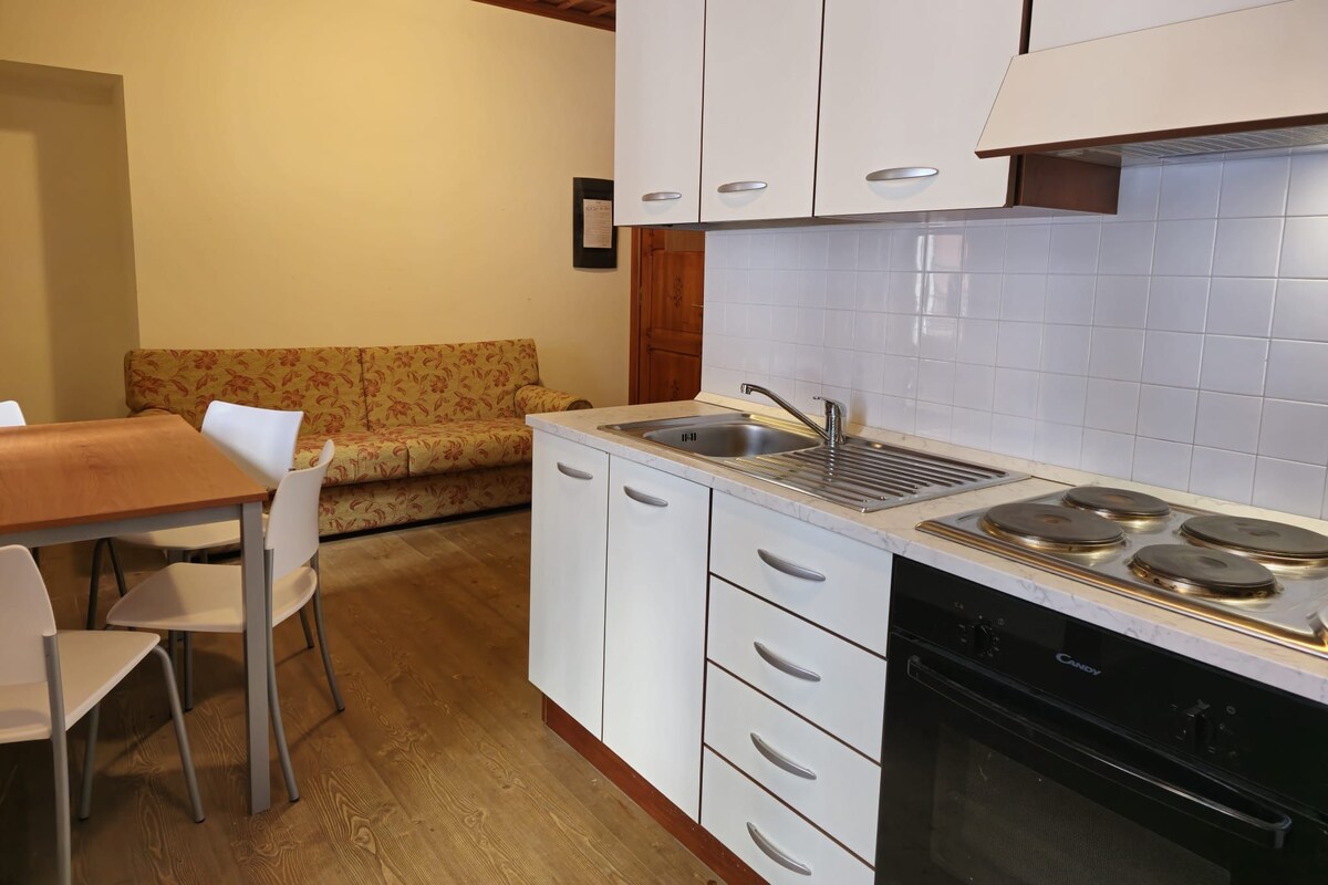 A compact kitchen with white cabinets, a built-in stove, and a sink. There is a wooden dining table with chairs and a floral sofa.