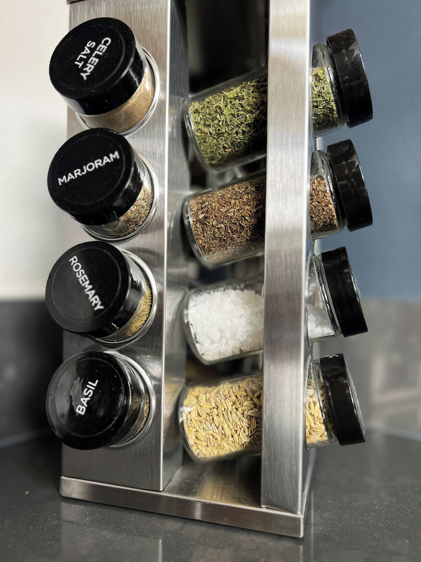 A spice rack with labeled containers for marjoram, rosemary, basil, and celery salt.