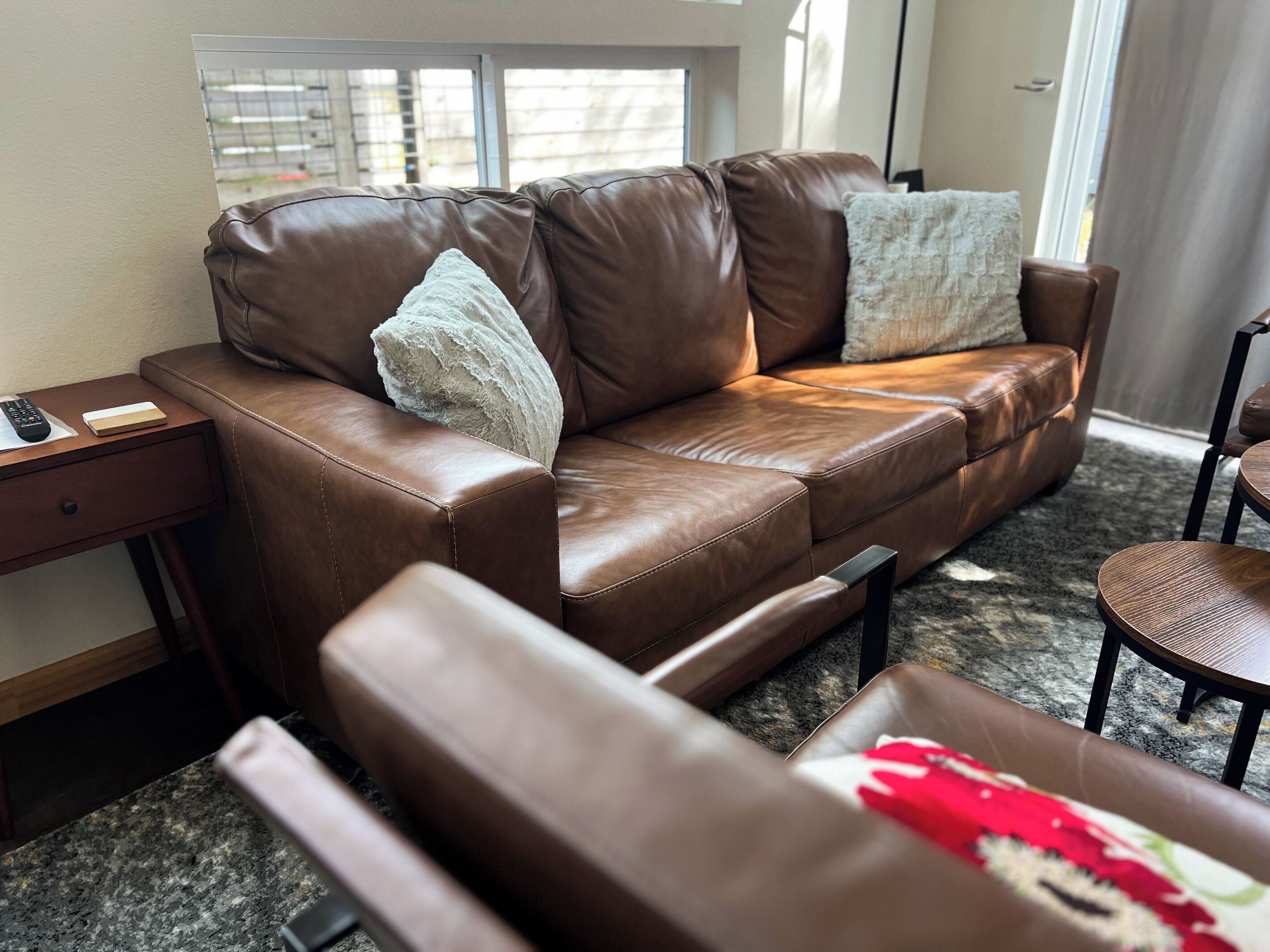 Un sofá de cuero marrón con dos almohadones, una mesita de madera con un control remoto y una silla de madera redonda.