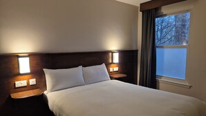 Habitació d'hotel amb un llit gran, dues taules de nit amb làmpades, una finestra amb cortines i vista a arbres a l'exterior.
