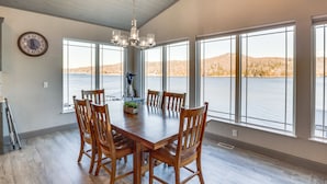 Un vaste espace dédié à la salle à manger, avec une grande table et des chaises en bois, de grandes fenêtres offrant une vue sur un lac et des collines, et un lustre suspendu au plafond.