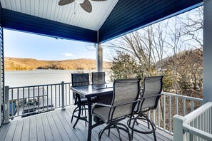 Een overdekt terras buiten met een tafel en stoelen, met uitzicht op een meer en heuvels.