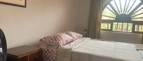 A bedroom with a bed, a desk, a chair, a standing fan, and a window with curtains.