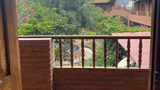 A balcony with a brick wall and a wooden railing overlooking a courtyard with greenery and a brick building.