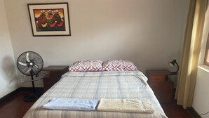 A neatly made bed with a plaid bedspread, two pillows with a geometric pattern, a standing fan, and a desk with a lamp.