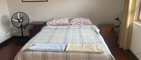 A neatly made bed with a plaid bedspread, two pillows with a geometric pattern, a standing fan, and a desk with a lamp.