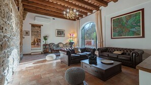 Un salon chaleureux avec un mur en pierre, des poutres apparentes en bois au plafond, un lustre, une grande fenêtre et un tableau.