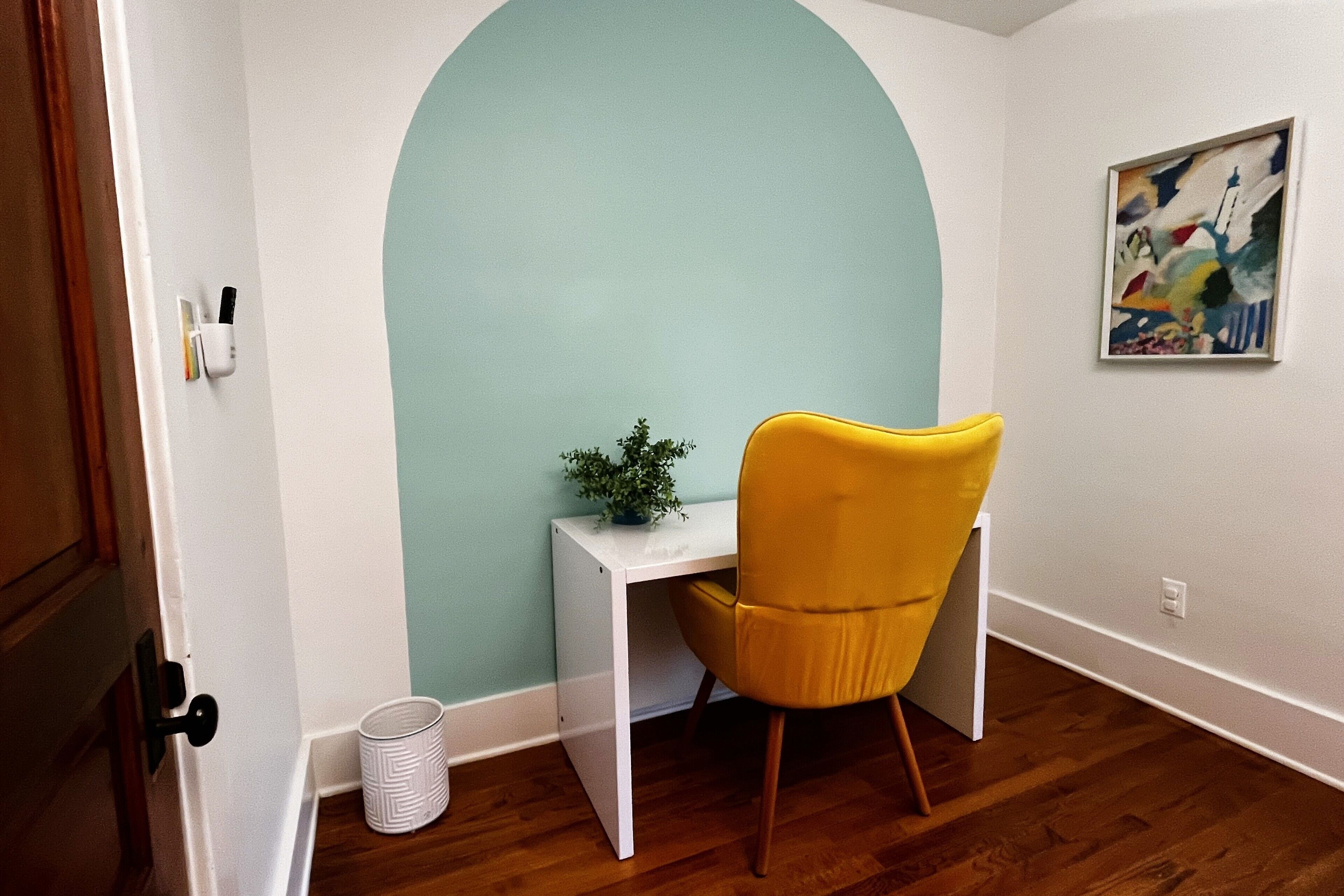 Ein Zimmer mit weißem Schreibtisch, gelbem Stuhl, Blumentopf und einem Bild an der Wand.
